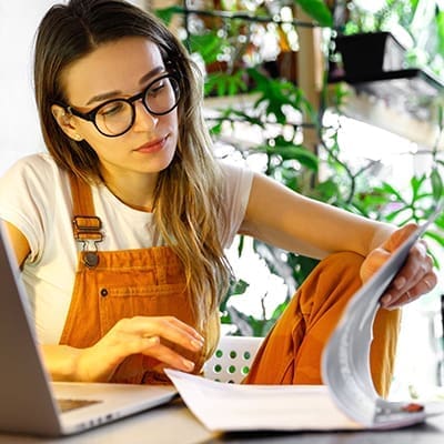 Young female gardener in glasses using laptop, makes a report on office documents, works from home during quarantine due to coronavirus. Cozy office workplace, remote work, E learning concept.
